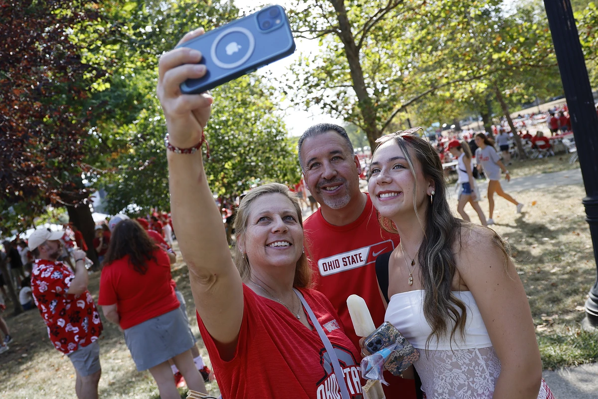 A group of three people are taking a selfie outdoors, wearing Ohio State attire in a festive setting.